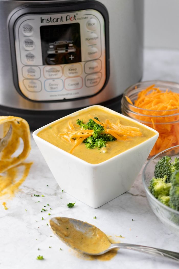 broccoli cheddar soup in a square bowl, topped with bits of broccoli, surrounded by the raw ingredients, and tools used to make it